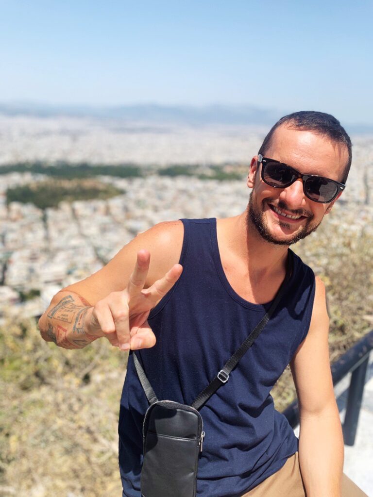 Chris in a blue tank top, with a black leather fanny pack wearing Rayban sunglasses and showing the peace sign with his fingers. The white buildings of Athens are behind him