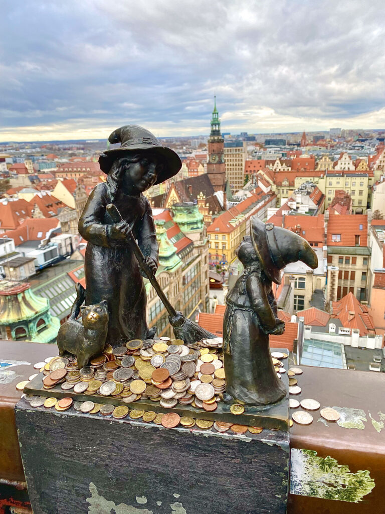 Top 7 Unique Travel Destinations to Visit in 2025 Close-up of two whimsical dwarf statues, part of Wroclaw, Poland's famous gnome trail, surrounded by coins left by visitors. The backdrop features the city’s charming red rooftops and iconic architecture, making Wroclaw a top pick for unique travel destinations.
